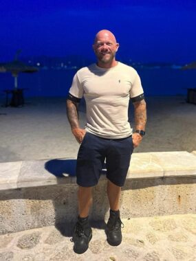 A man poses for a photo in front of the beach at dusk. He's wearing a white T-shirt, dark shorts, and black sneakers and has the muscular physique of a bodybuilder.