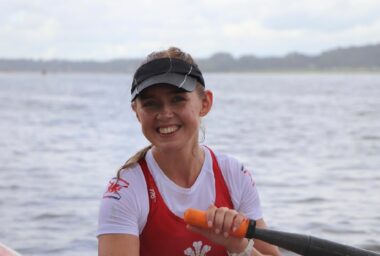 A young woman smiles in front of a large body of water. She's wearing a black visor and a red and white rowing uniform, and is holding an oar with her left hand. The photo is cropped below her chest, but we can assume she's sitting in a rowboat.