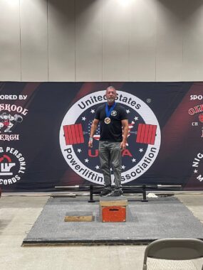 A man stands atop a wooden box in front of a barbell. A large banner behind him reads "United States Powerlifting Association." He is standing proudly and is wearing a medal around his neck.