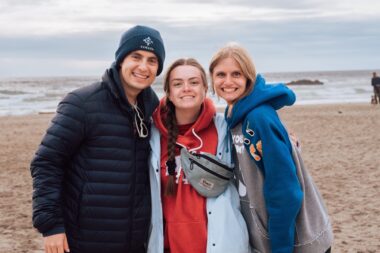Three young adults wearing hoodies and jackets stand side by side on a beach. It looks like a cloudy, chilly day, but all three are smiling happily.