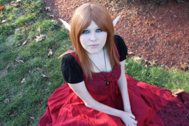 A young woman is dressed in a cosplay outfit that includes a red dress and pointed ears. She's seated on the grass with her hands in her lap and looking up at the camera.