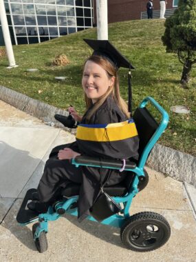 A young woman wearing a graduation cap and gown is seated in a power wheelchair with a turquoise frame. She's on an outdoor walkway on what appears to be a college campus and is smiling at the camera to her left.